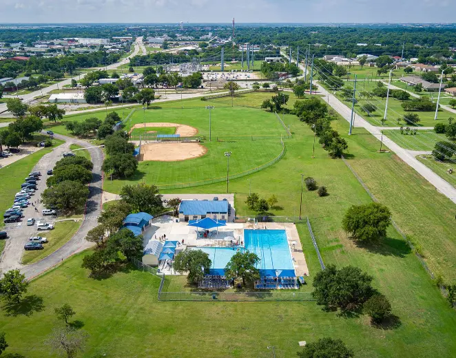 Aerial view of community park with swimming pools, baseball diamonds, and expansive green fields surrounded by trees and parking areas