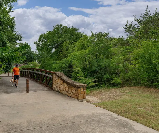 Runner jogging on concrete path beside wooden bridge rail with trees and grassy area