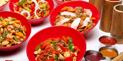 Multiple red bowls filled with Mongolian stir-fry containing meat, vegetables, and noodles, with sauce bowls nearby