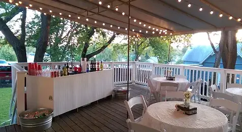 Outdoor covered bar setup with white railings, round table, chairs, and string lights overhead in a grassy venue