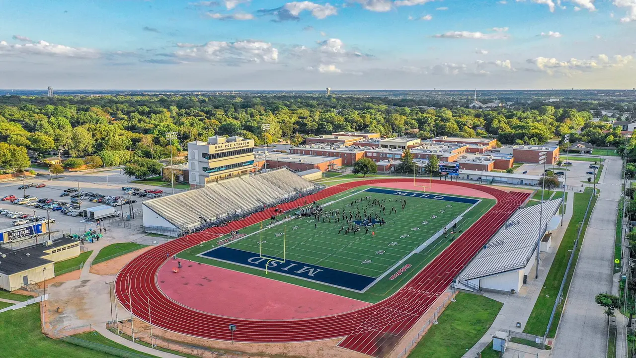 Aerial view of athletic stadium with red running track, green field, surrounding school buildings and wooded landscape under partly cloudy sky