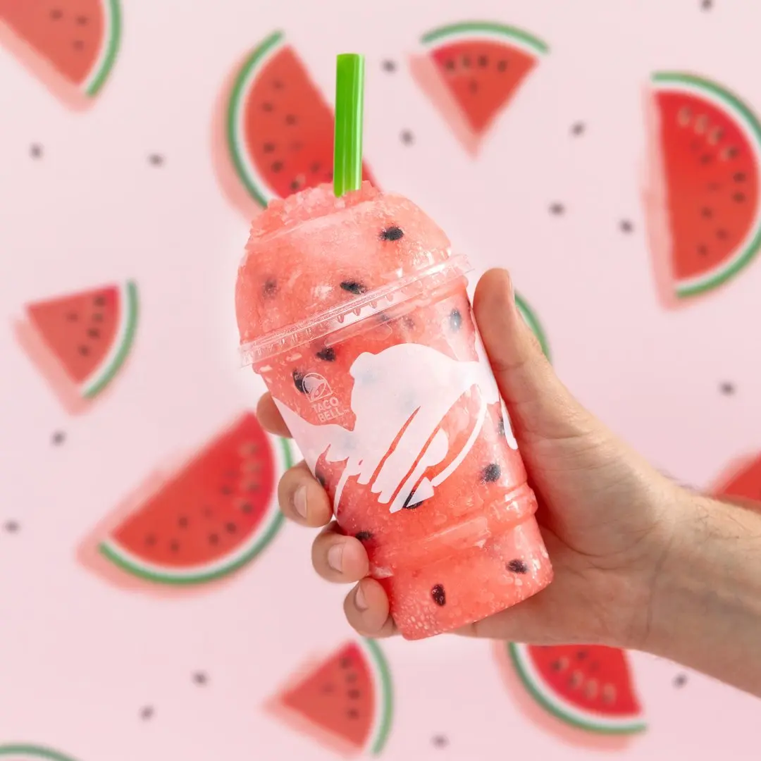 Hand holding a pink watermelon-themed frozen drink with green straw against pink background with watermelon slice graphics