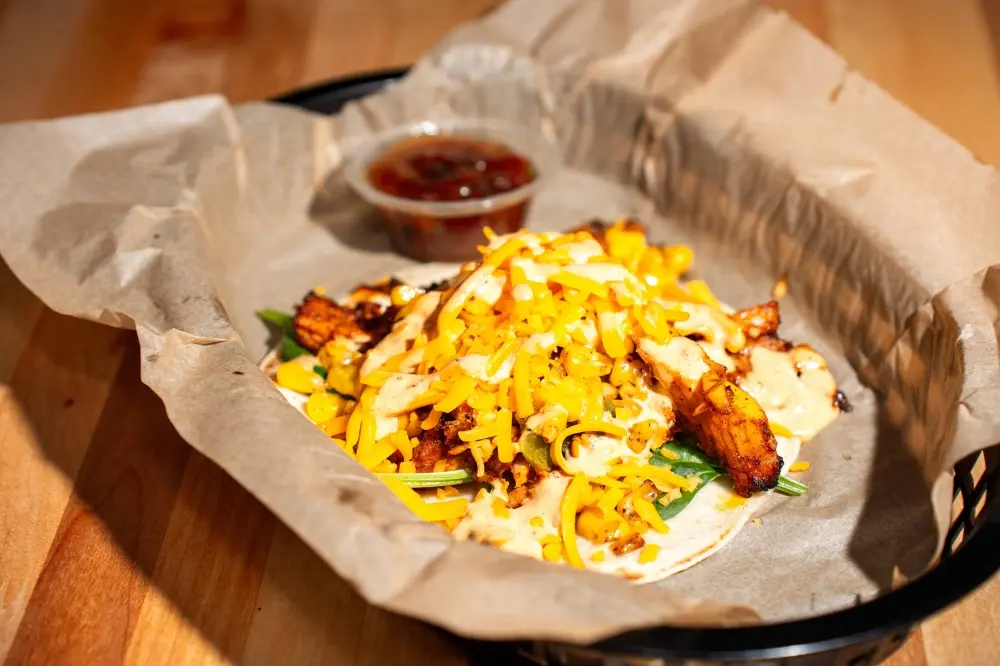 Shredded cheese and crispy bacon over eggs and greens in a paper-lined container with sauce on the side