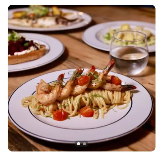 Shrimp and pasta dish with red peppers on a white plate, alongside other plated meals and water glasses on a wooden table