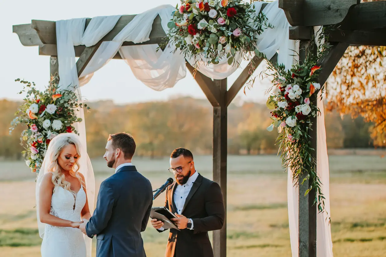Bride and groom exchanging vows under flower-adorned wooden pergola with officiant, autumn landscape visible behind them