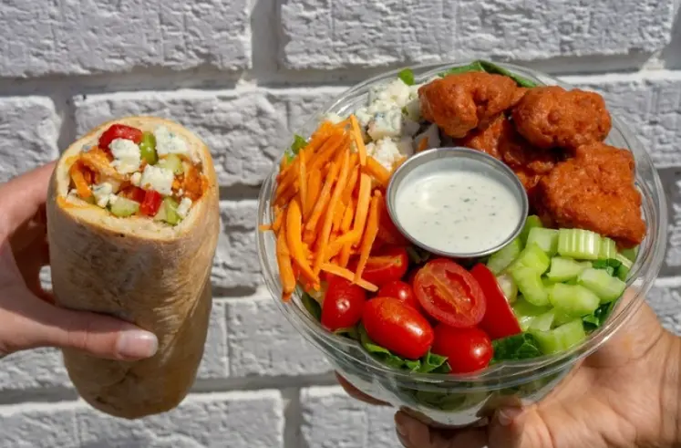 Hand holding wrapped sandwich and platter with fried chicken, french fries, fresh vegetables, and dipping sauce against white brick wall