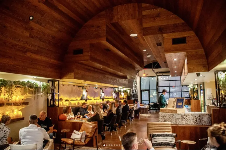 Warm wood-beamed interior of a wine bar with customers seated at tables and a bar counter, ambient lighting creating a cozy atmosphere