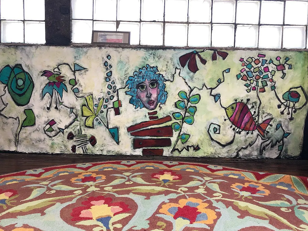 Colorful mural with whimsical figures and plants on cream wall above patterned red and gold rug in indoor space