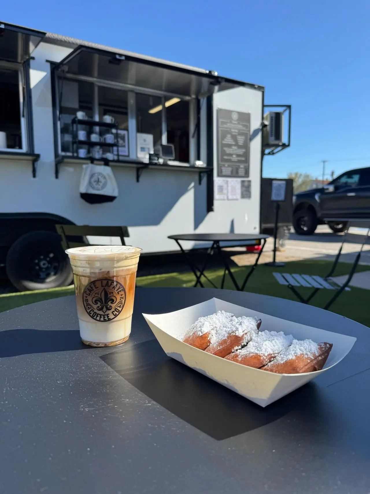 Iced coffee drink and powdered sugar-dusted beignets on outdoor table in front of food trailer