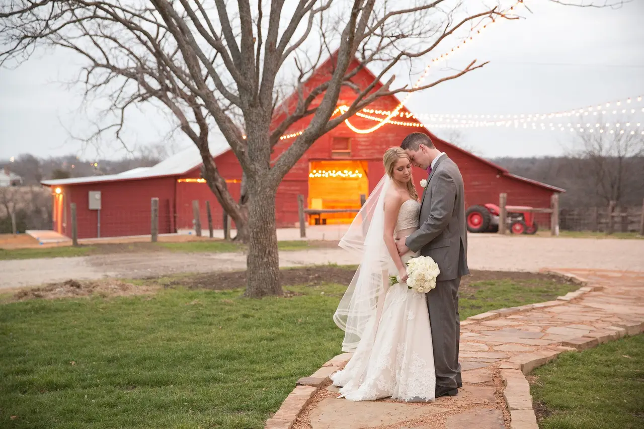 Bride and groom kissing under bare tree in front of red barn with brick pathway