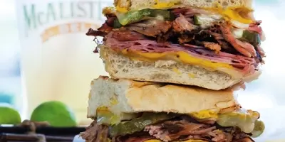 Stacked sandwich with roast beef, cheese, and vegetables on bread, served with a beverage and lime