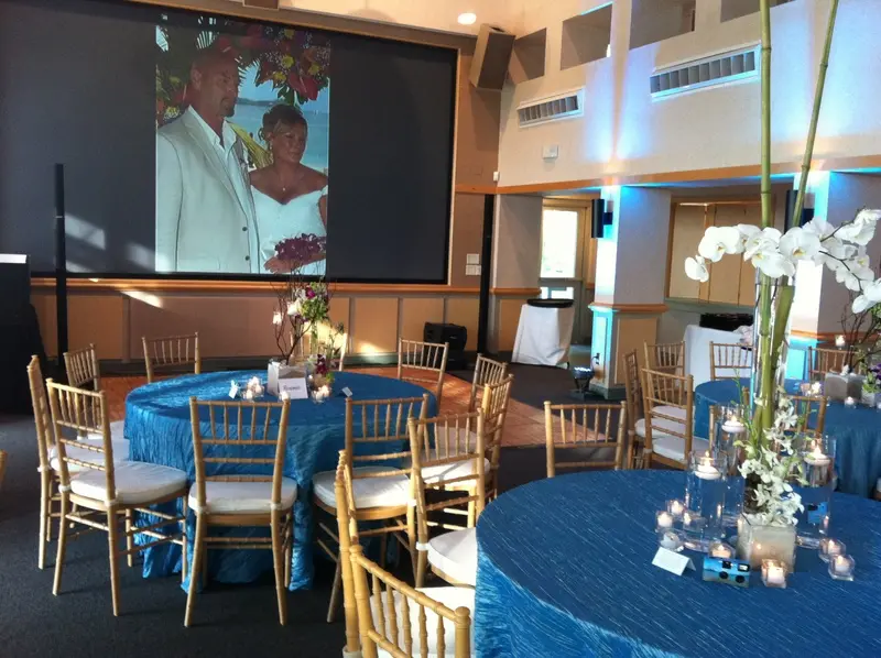 Event space with round tables dressed in blue linens, gold chairs, white floral centerpieces, and projection screen displaying a couple's photo