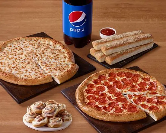 Two pizzas with toppings, breadsticks, cinnamon rolls, sauce cup, and bottle of Pepsi arranged on wooden surface