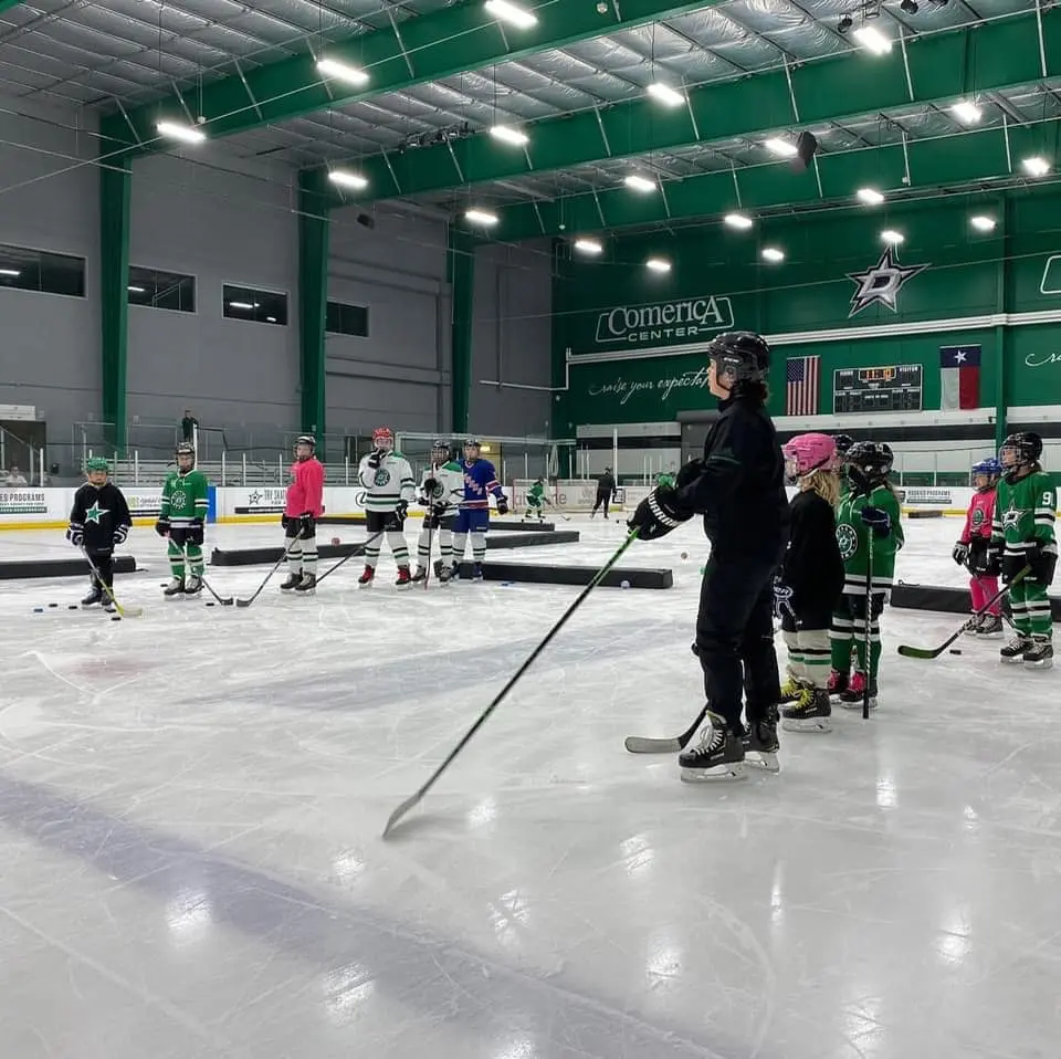 An adult instructor demonstrates hockey technique to young players on an indoor ice rink during a training session
