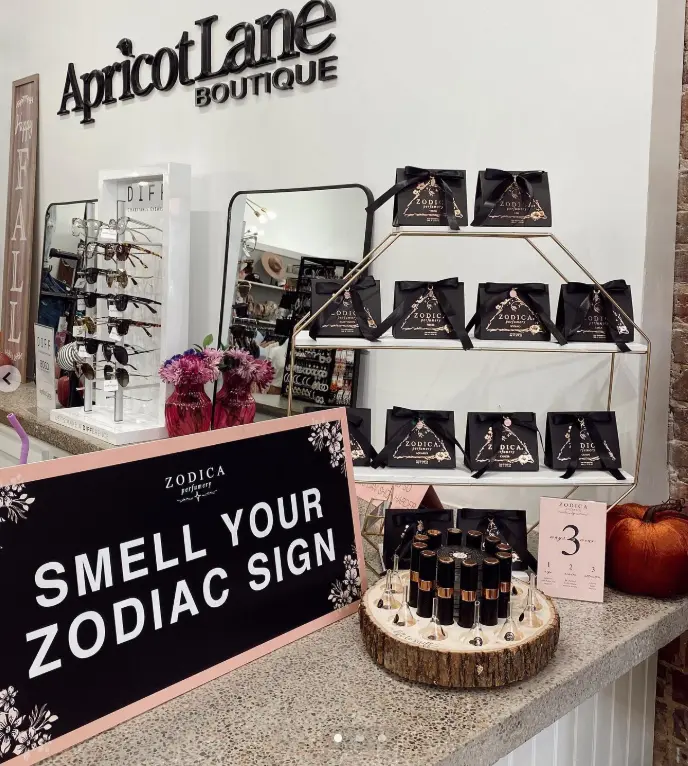 Boutique counter display with zodiac-themed candles, pink flowers, mirror, and "Smell Your Zodiac Sign" signage