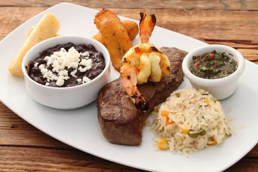 Grilled steak with skewered shrimp, black beans with cheese, rice with vegetables, plantain chips, and chimichurri sauce on white plate