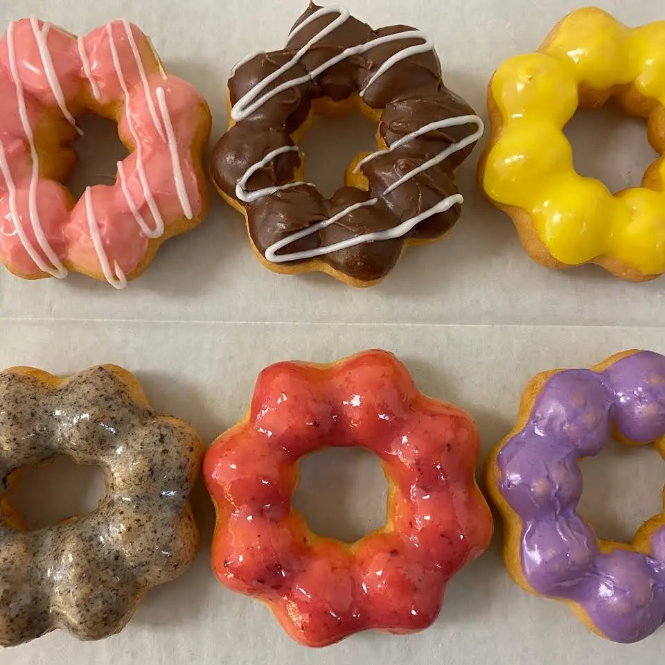 Six frosted donuts with various toppings: pink glazed with white drizzle, chocolate with white drizzle, yellow glazed, speckled brown, red glazed, and purple