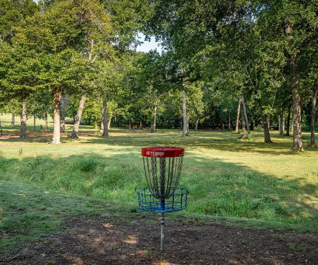 Red and blue disc golf basket on a tree-lined grass course surrounded by mature oaks