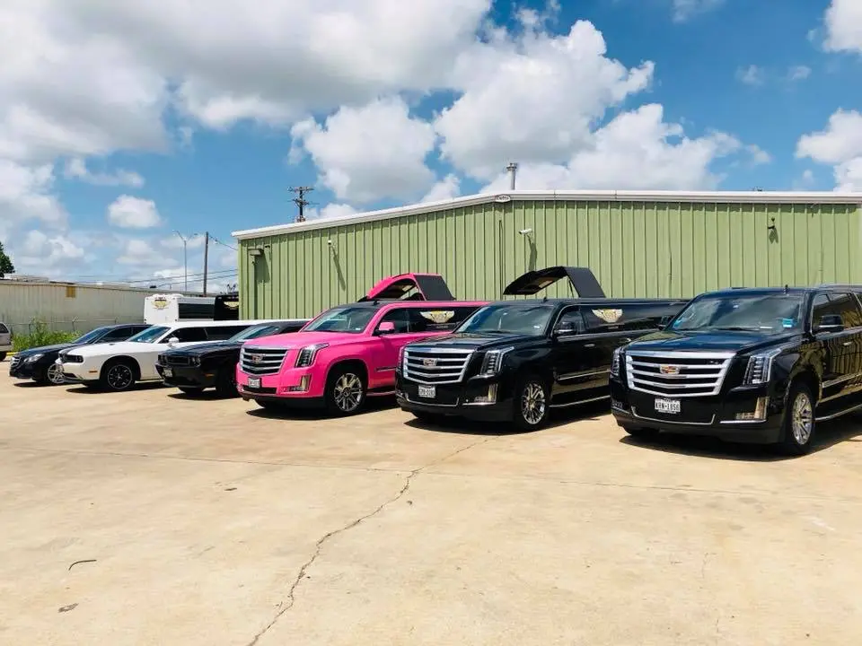 Fleet of luxury vehicles including pink and black SUVs and sedans parked on concrete lot in front of green metal warehouse building