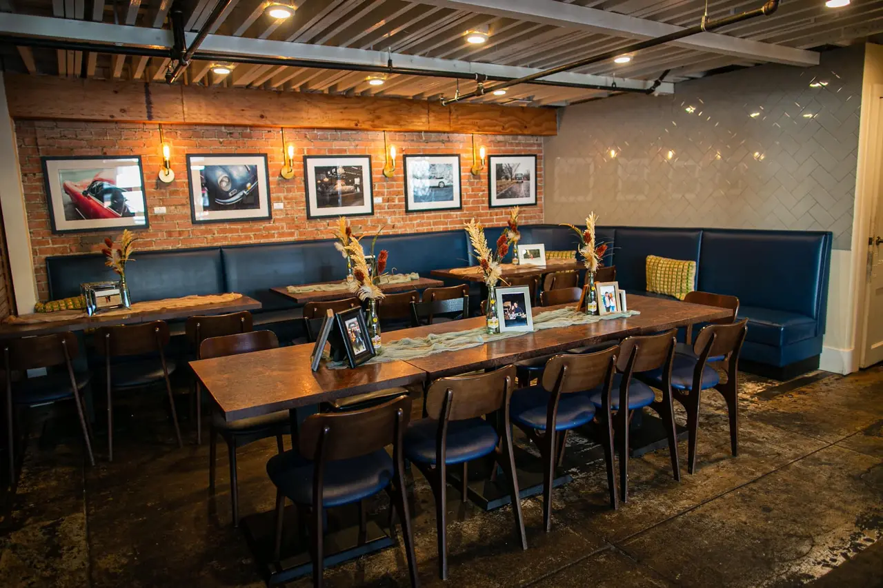 Interior bar and event space with wooden tables, blue upholstered seating, framed photographs on brick wall, and industrial ceiling