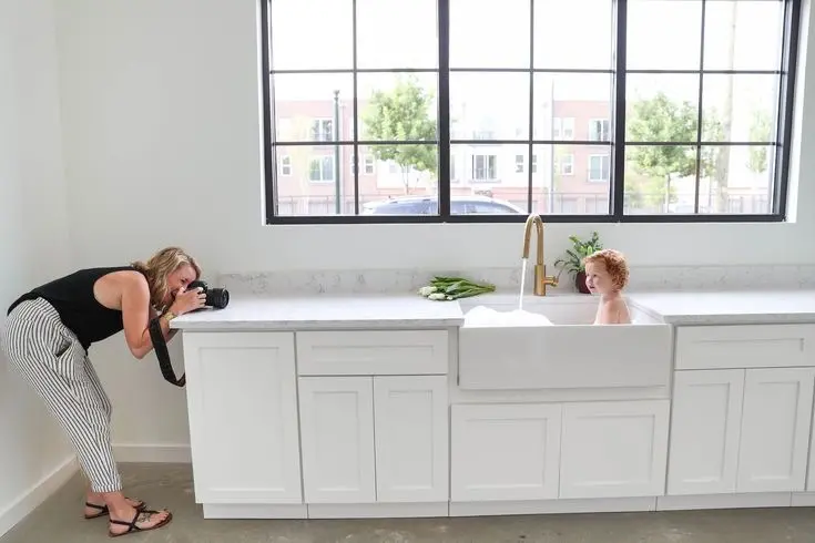 Woman leaning over white kitchen counter with large windows behind, potted plant on counter