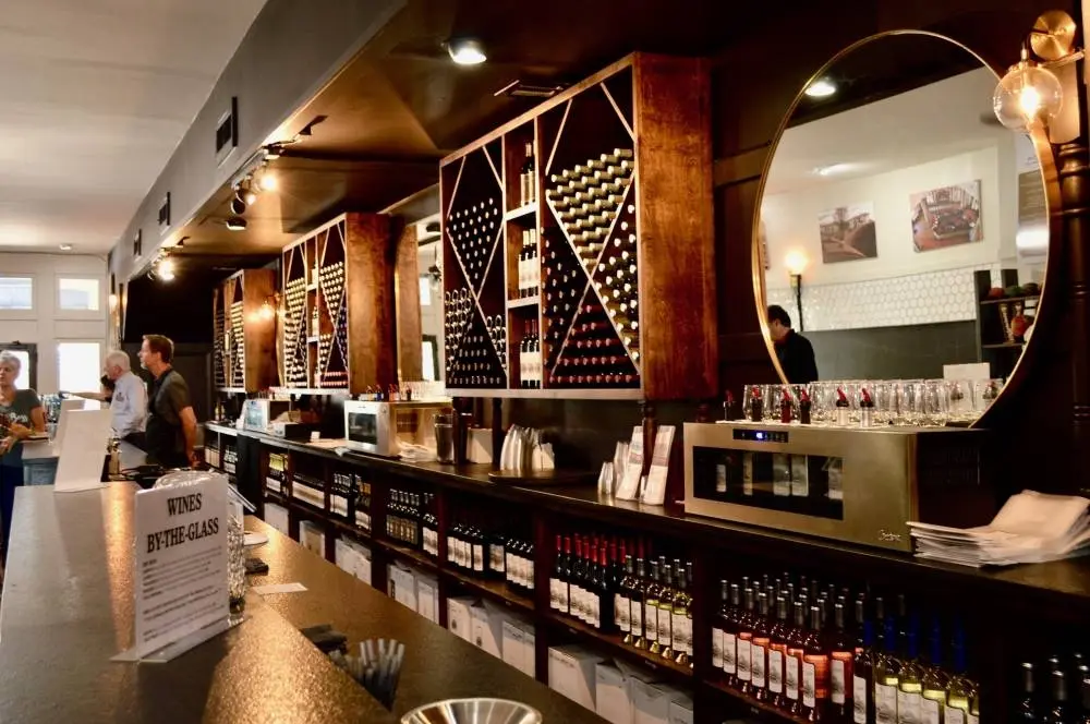 Wine tasting room with dark wood counter, wine bottle displays, and customers browsing merchandise under warm lighting