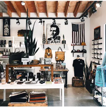Modern retail shop interior with wooden beams, merchandise displays on white walls, black posters, plants, clothing racks, and shelving with curated goods