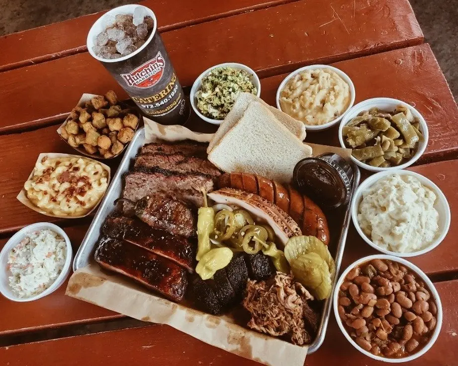 Overhead view of barbecue platter with brisket, ribs, sausage, pickles, white bread, and assorted side dishes in bowls on wooden table