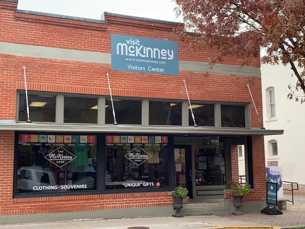 Brick storefront with "visit mckinney" signage above large windows displaying clothing and souvenirs, with planters flanking the entrance