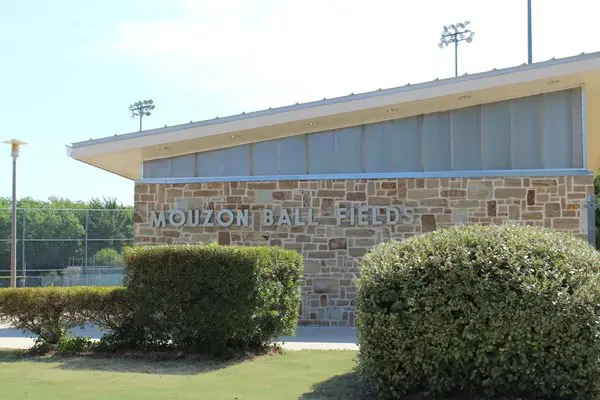 Modern sports facility building with stone exterior, manicured landscaping, and stadium lighting towers visible behind