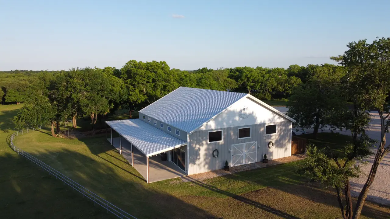 White ranch building with metal roof and covered porch surrounded by green fields and scattered trees, aerial view