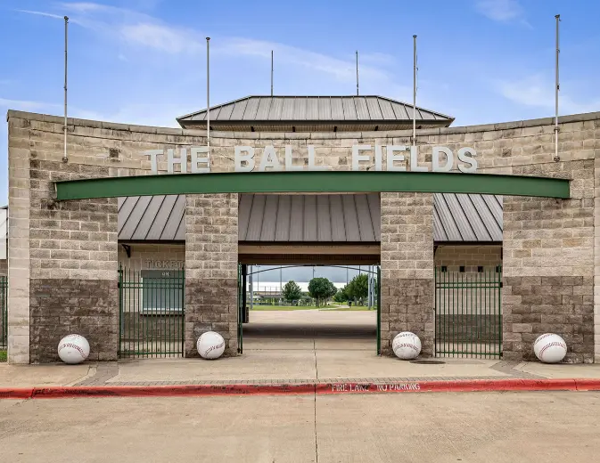 Brick entrance building with green awning and large opening framing baseball fields beyond, flanked by decorative spheres
