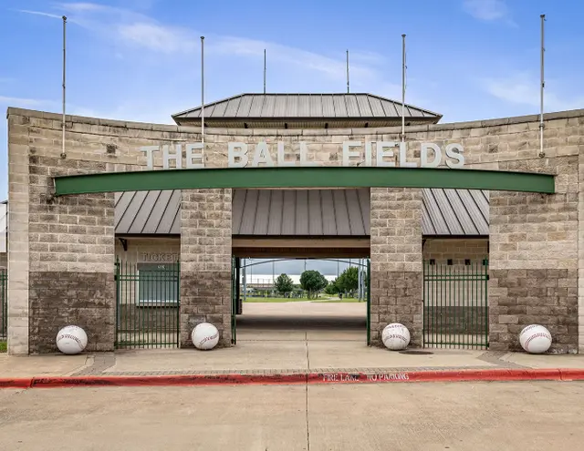 Brick entrance building with green awning and large opening framing baseball fields beyond, flanked by decorative spheres