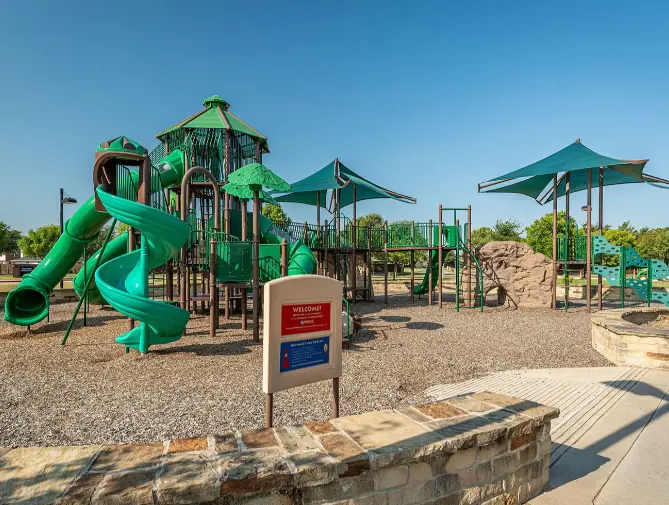 Playground with bright green spiral slides and climbing structures, shaded by teal canopies, on wood chips with clear blue sky