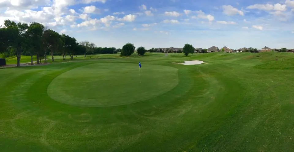 Manicured golf course green with fairway, flag marker, trees lining the perimeter, and residential buildings visible in the distance under blue sky
