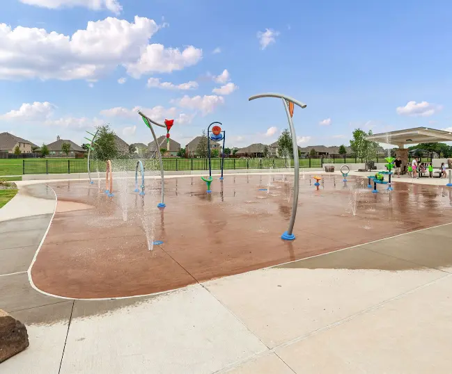 Splash pad with spray features and water jets on a sunny day, surrounded by residential neighborhood and trees
