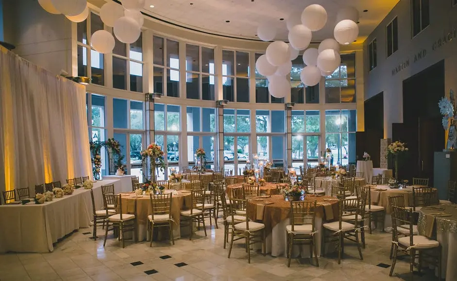 Event venue with round tables, wooden chairs, white paper lantern decorations, and floor-to-ceiling windows overlooking the city