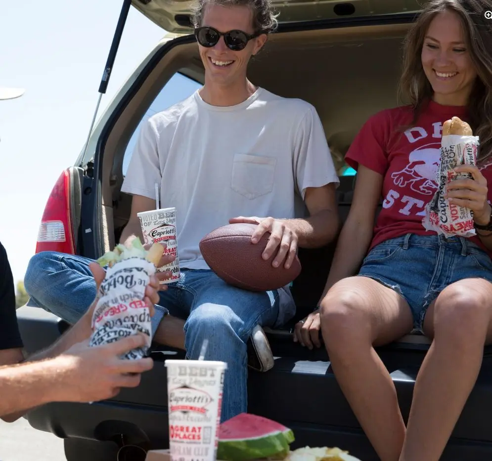 Three people sitting in open car trunk sharing food and drinks on sunny day