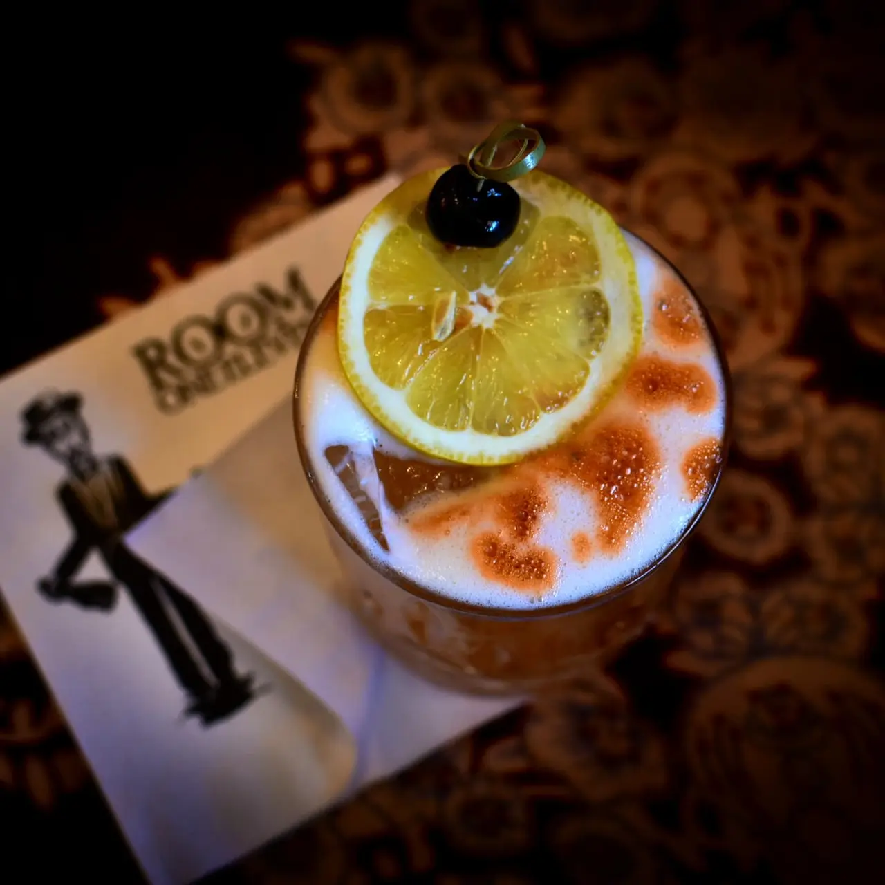 Cocktail with lemon slice garnish and dark cherry, topped with cinnamon-dusted foam, served beside Room One Eleven napkin