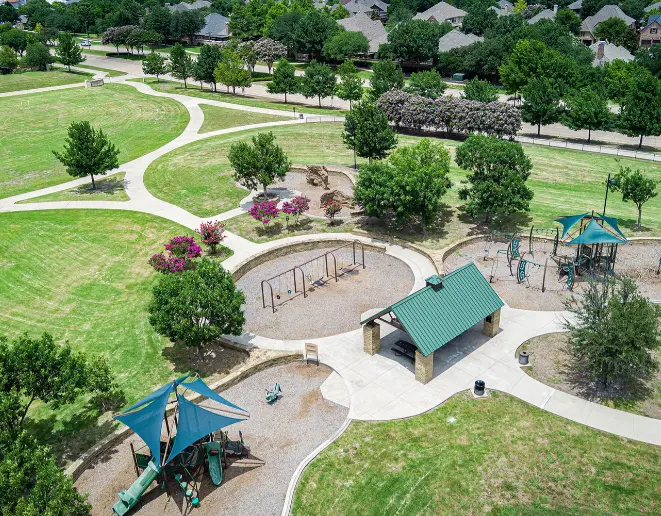 Aerial view of neighborhood park with playground equipment, open lawns, walking paths, shade trees, and residential homes beyond