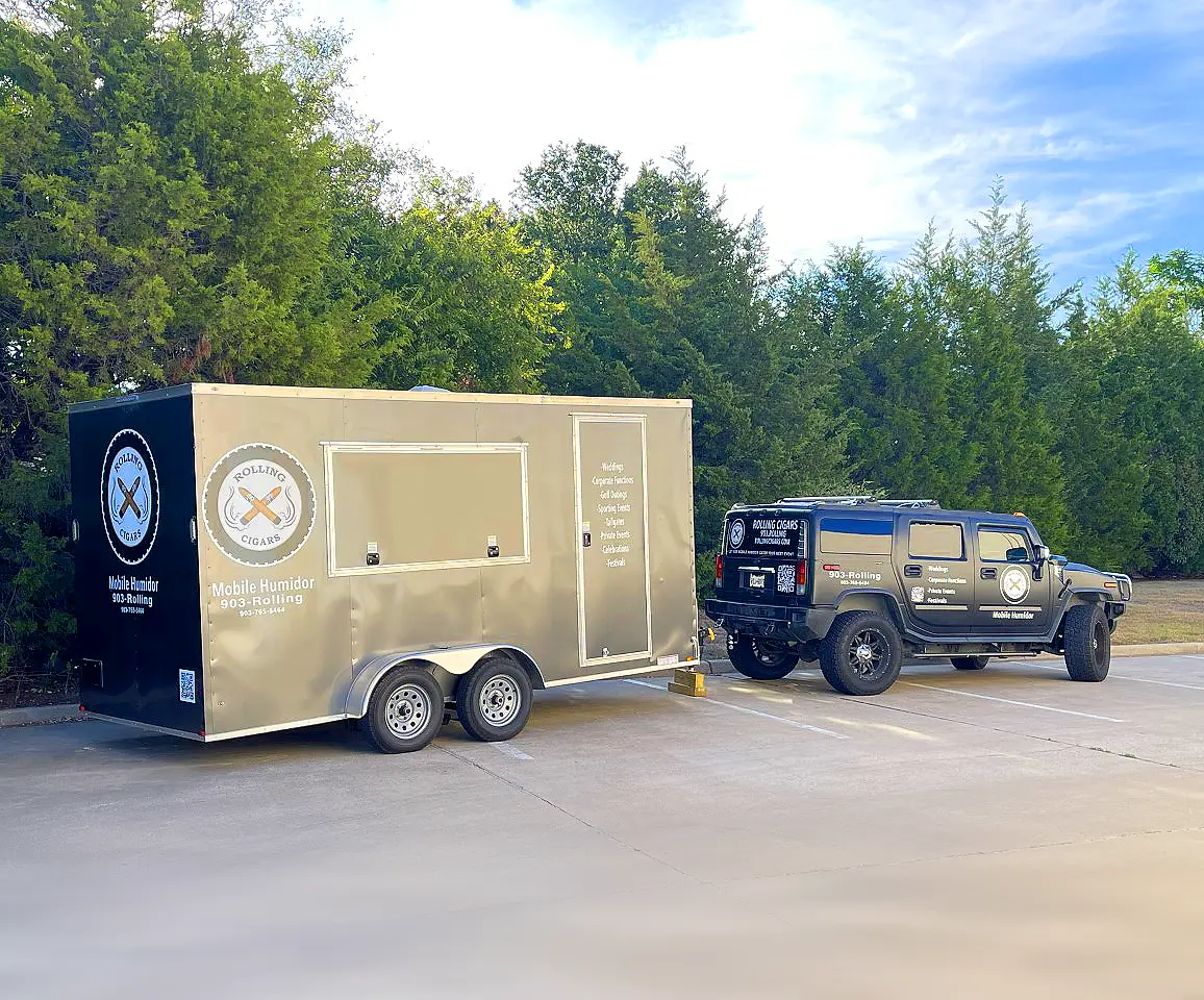 Tan mobile cigar humidor trailer parked next to a dark vehicle in a lot, with trees in background