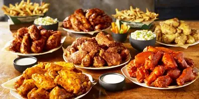 Platters of sauced chicken wings in various flavors arranged with sides of french fries and dipping sauces on a wooden table