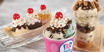 Layered ice cream sundae in pink cup with whipped cream, chocolate sauce and cherry on top, alongside ice cream cones and parfait in clear cup