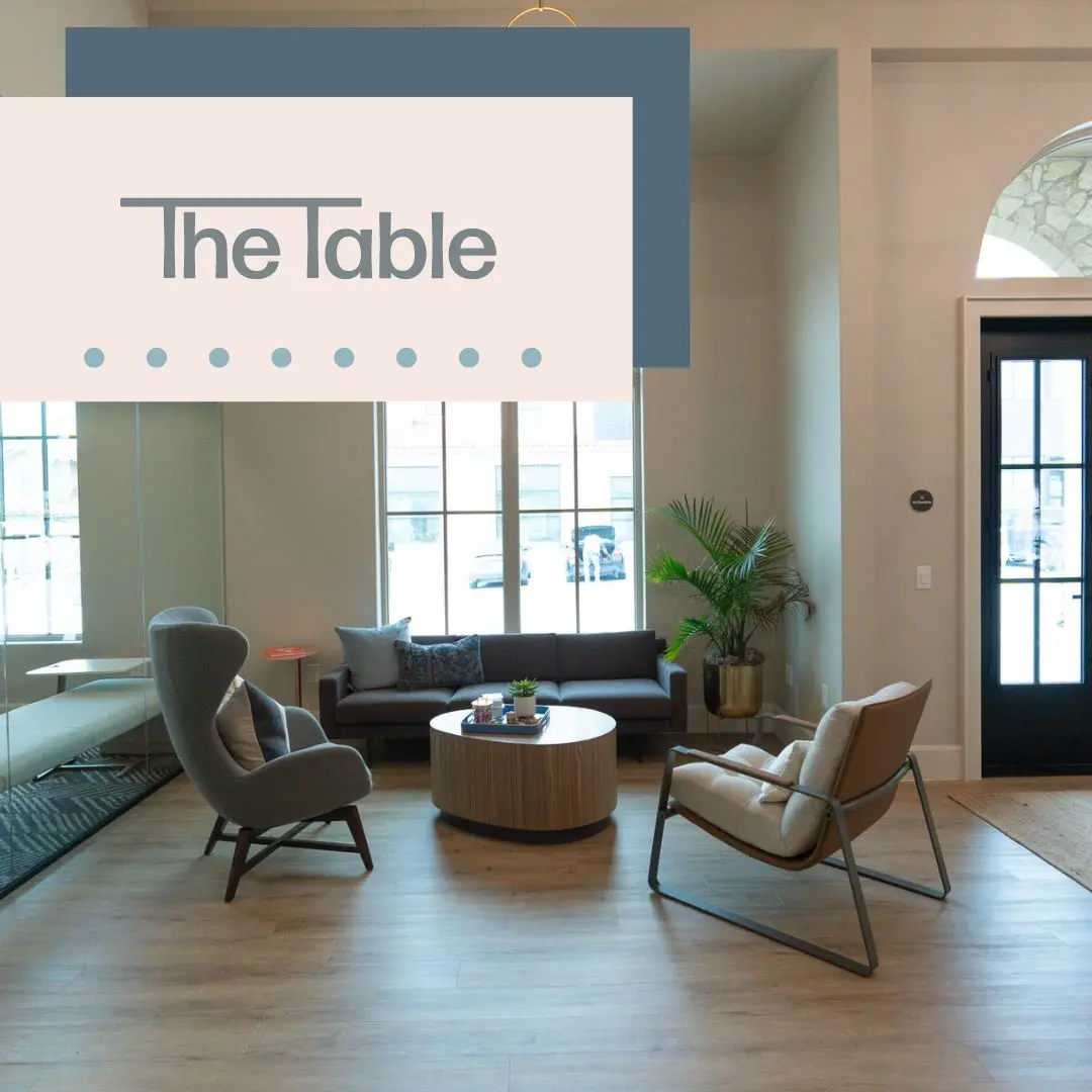 Modern furniture showroom with two upholstered chairs facing a round wooden table, sofa and windows in background
