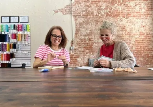 Two women working at a wooden counter in a craft shop with exposed brick walls and colorful yarn displays