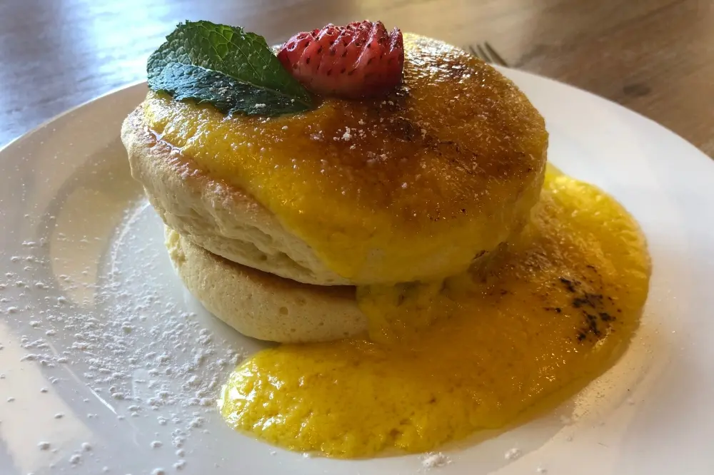Stack of fluffy pancakes topped with mint leaf and raspberry, served with yellow sauce on a white plate