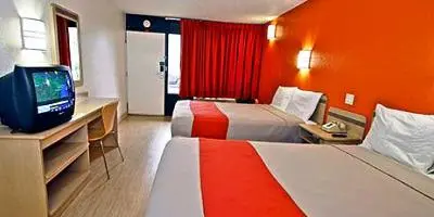 Hotel room with two beds with red runners, orange accent wall, red curtains, and desk with television