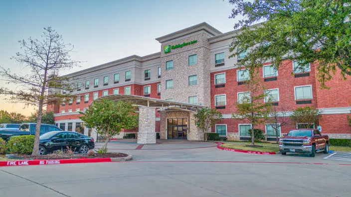 Red brick hotel building with covered entrance, parking lot, and surrounding landscaping at dusk