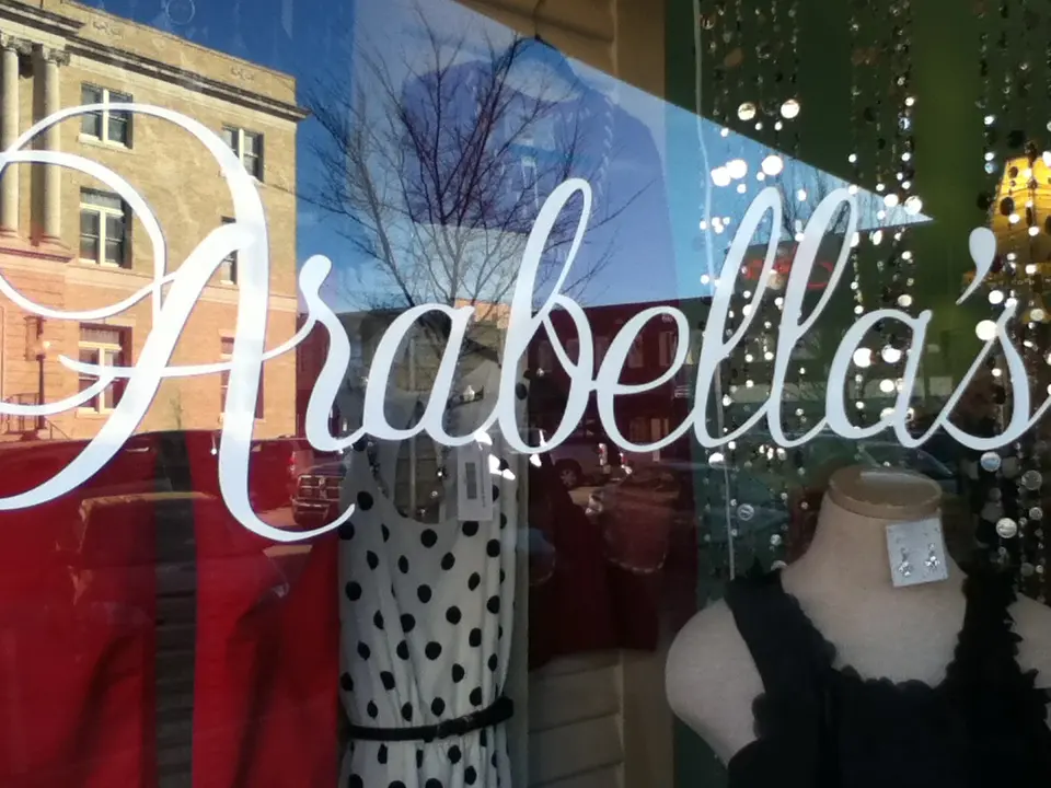 Store window display with mannequins wearing clothing and decorative items behind glass storefront