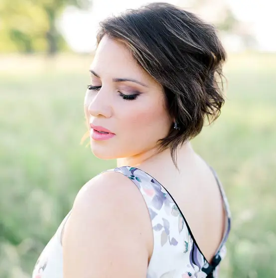 Woman with styled dark hair and pink lipstick, looking downward outdoors with blurred green background